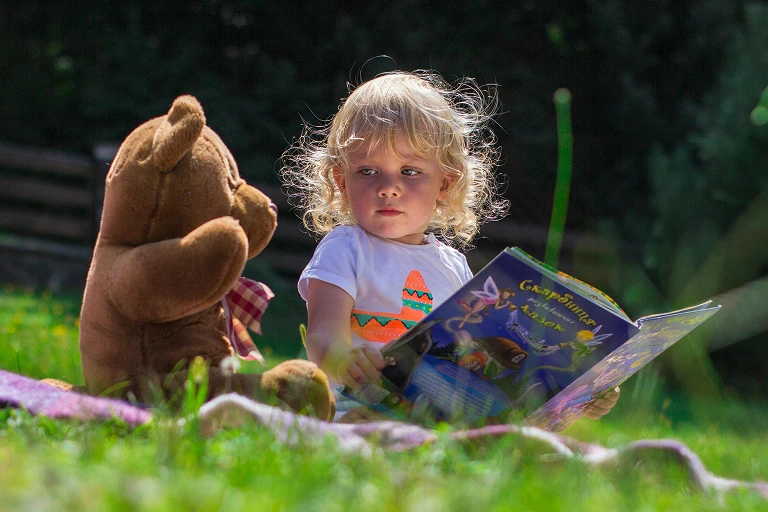Young child sitting outdoors and looking at a picture book beside a soft toy