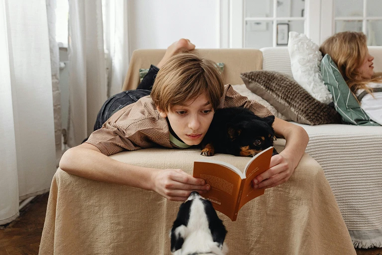 Child lying on a sofa and reading a book with a pet nearby