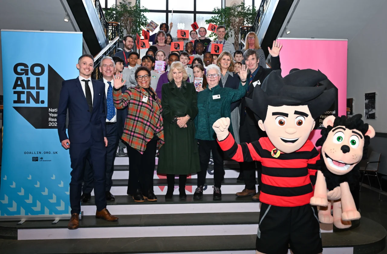 A photograph showing Her Majesty the Queen amongst a group of adults and school children standing on a staircase at the National Library of Scotland, accompanied by Dennis the Menace and Gnasher.