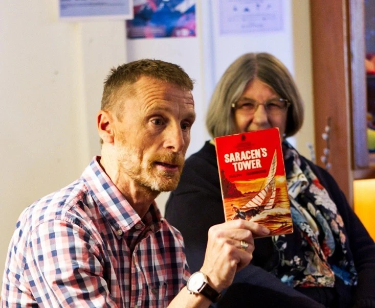 A main holding up a book with a woman smiling, looking torwards him in the background