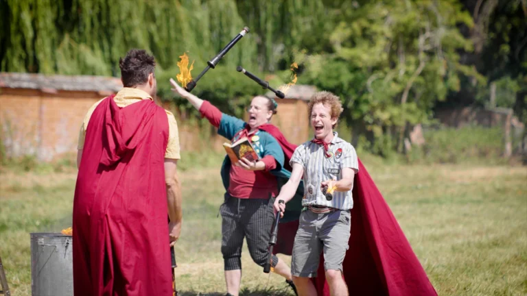 Two adults looking very animated, wearing capes running outside towards a third adult in a cape. One of the adults appears to be reading a book whilst the other is juggling lit torches. Part of an outdoor arts festival.